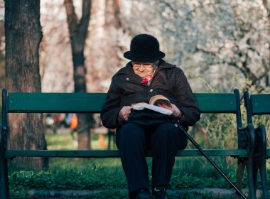 person sits on bench reading book in front of tree at daytime