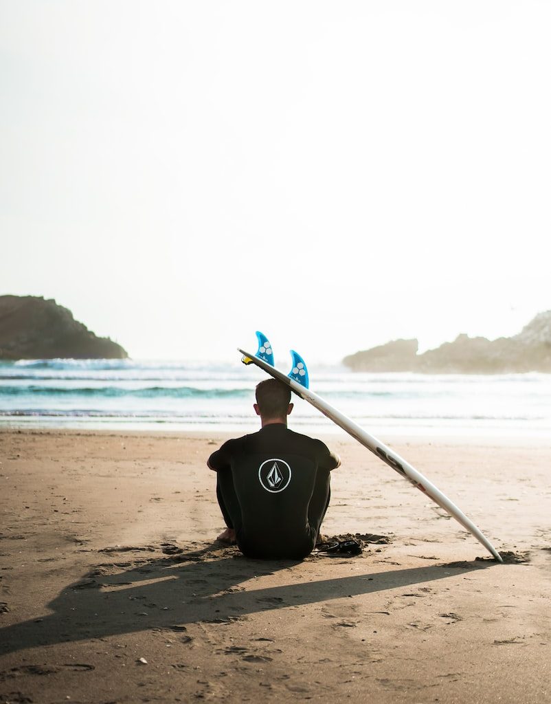 man sitting on sand beside surfboard facing sea during daytime