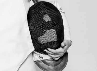 person wearing white fencing suit holding black mask inside room