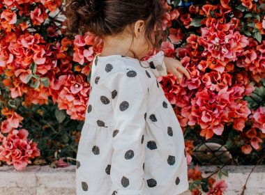 girl in black and white polka-dot long-sleeved blouse and blue shorts standing beside red petaled flowers during daytime