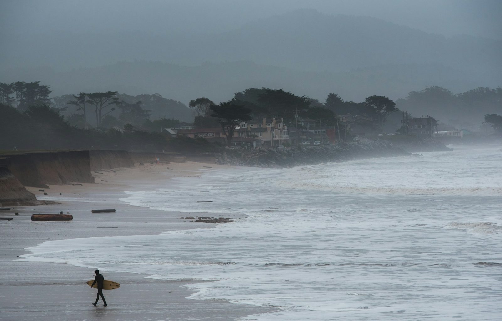 person walking and carrying surfboard on seashore during foggy weather