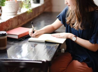 woman sitting in front of black table writing on white book near window