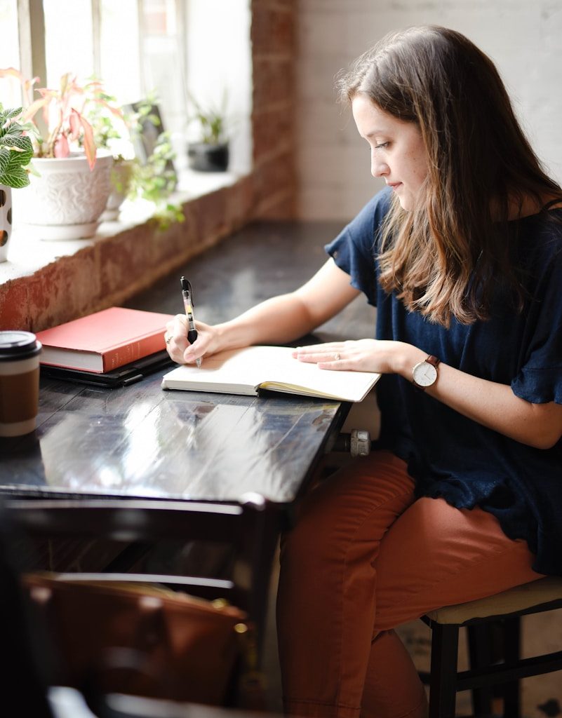 woman sitting in front of black table writing on white book near window