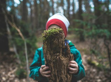 child in blue jacket and white knit cap holding brown tree trunk