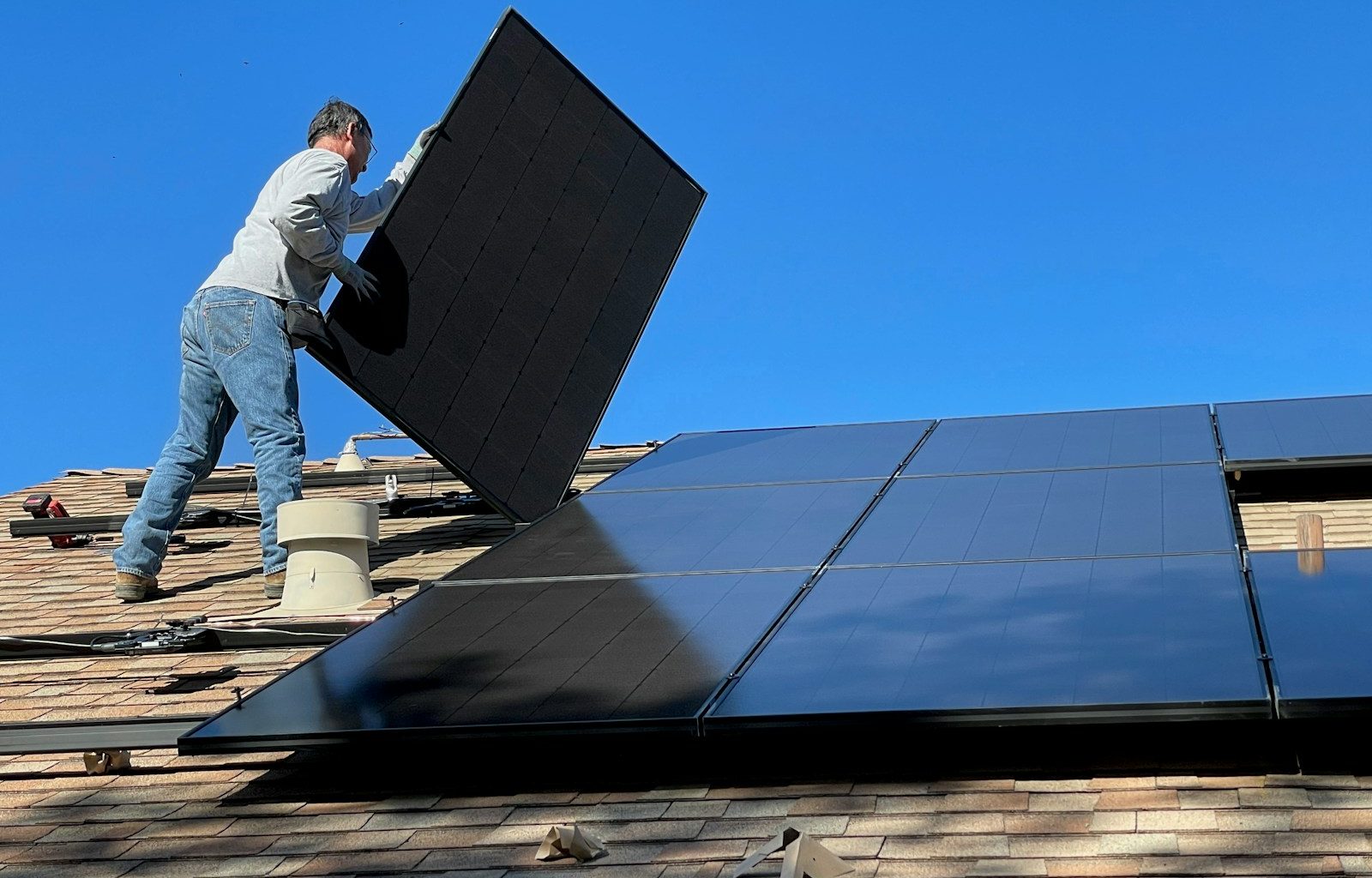 A person installing a black solar panel on a shingled roof under blue sky