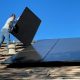 A person installing a black solar panel on a shingled roof under blue sky